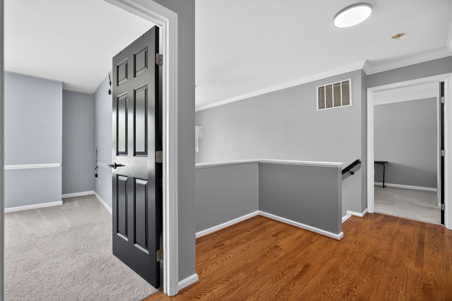 Modern home interior hallway featuring hardwood floors, gray walls, black six-panel door, and open staircase railing.
