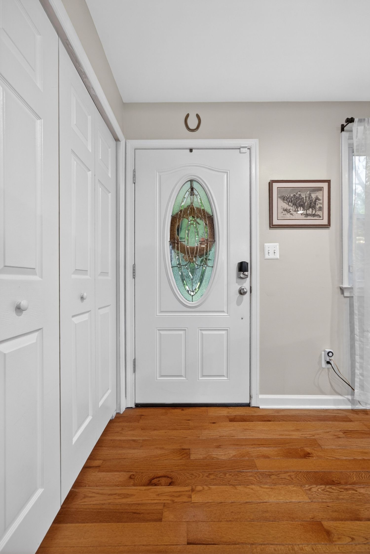 Bright entryway with hardwood floors, white front door featuring oval stained glass insert, and adjacent white closet doors.