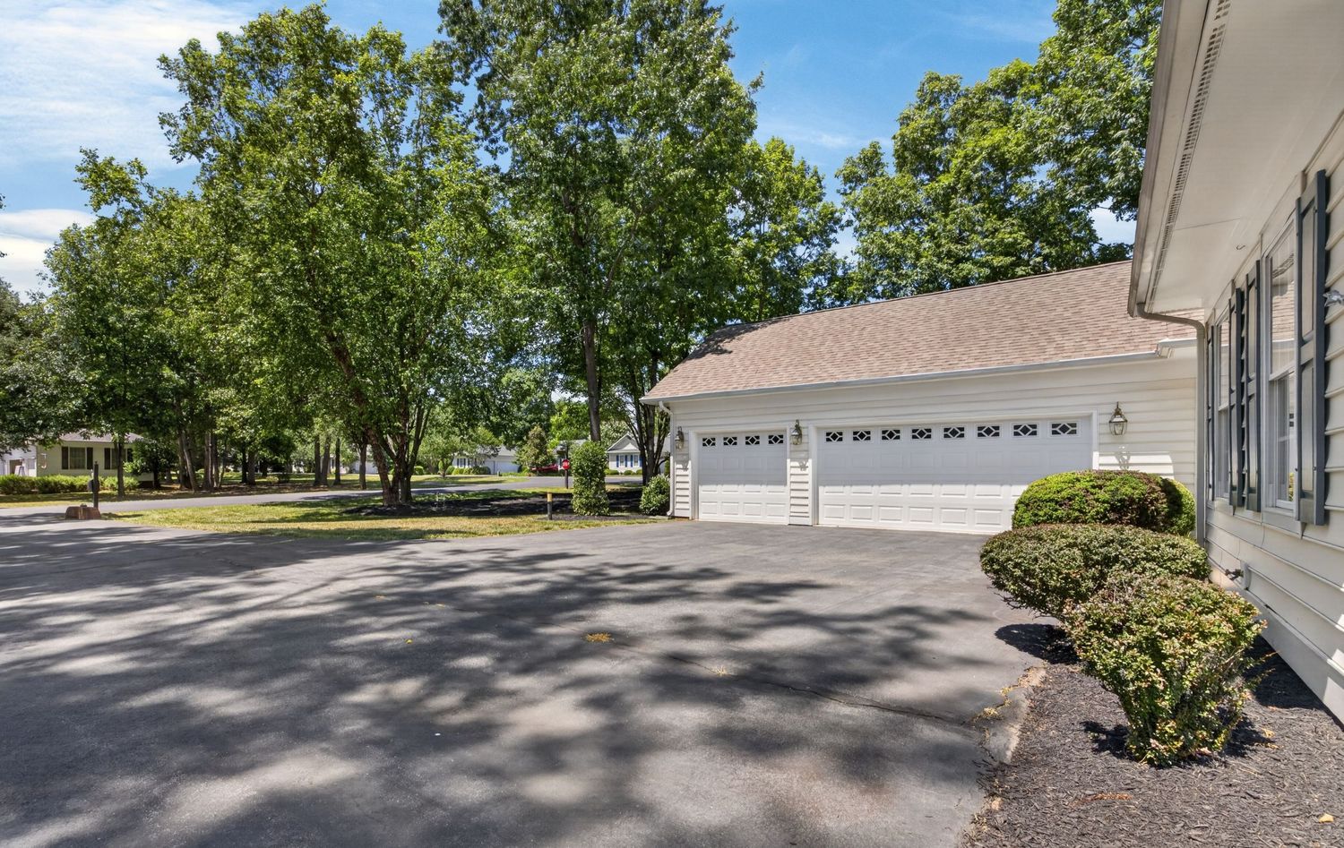 White two-car garage beside a home surrounded by lush green trees on a sunny day with a paved driveway.