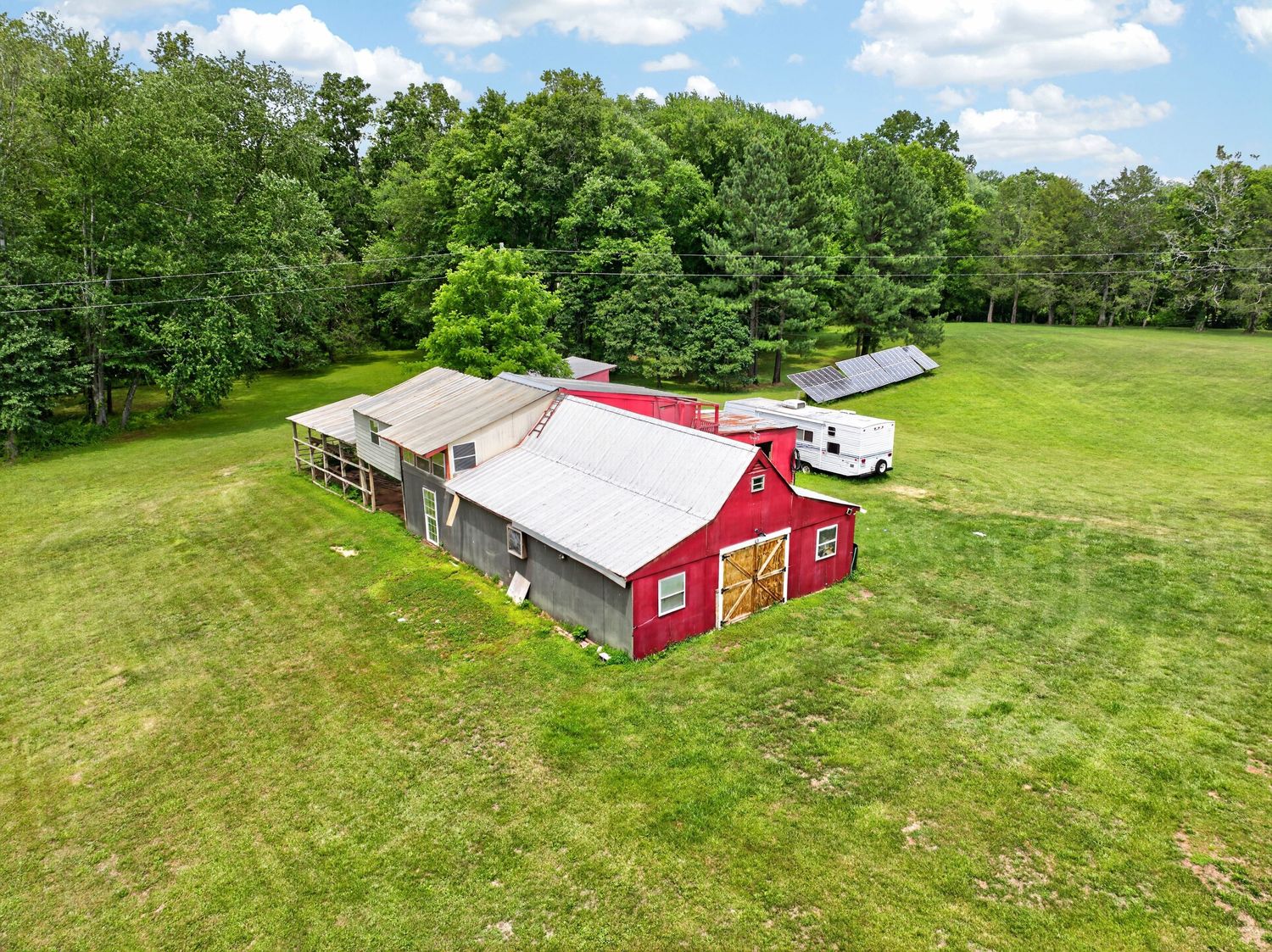 Aerial view of a red barn with metal roof surrounded by lush green fields and dense trees on a sunny day.