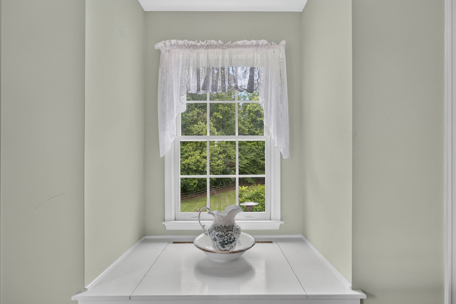 White-framed window with sheer curtains overlooking lush green trees, above a white ledge with a decorative teacup.