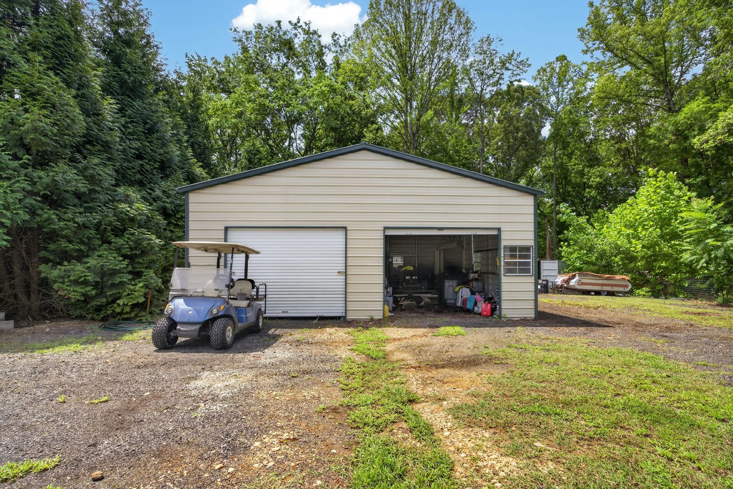 A golf cart parked outside a beige metal garage with an open bay, surrounded by lush green trees.
