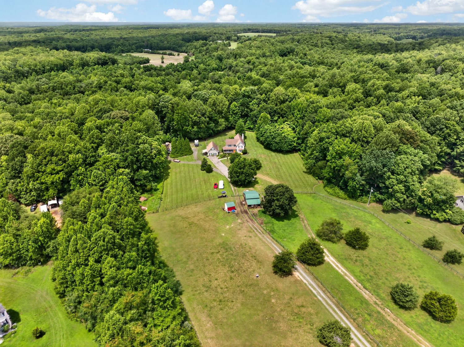Aerial view of a lush green rural property with farmland, scattered buildings, and dense surrounding woodland under a blue sky.