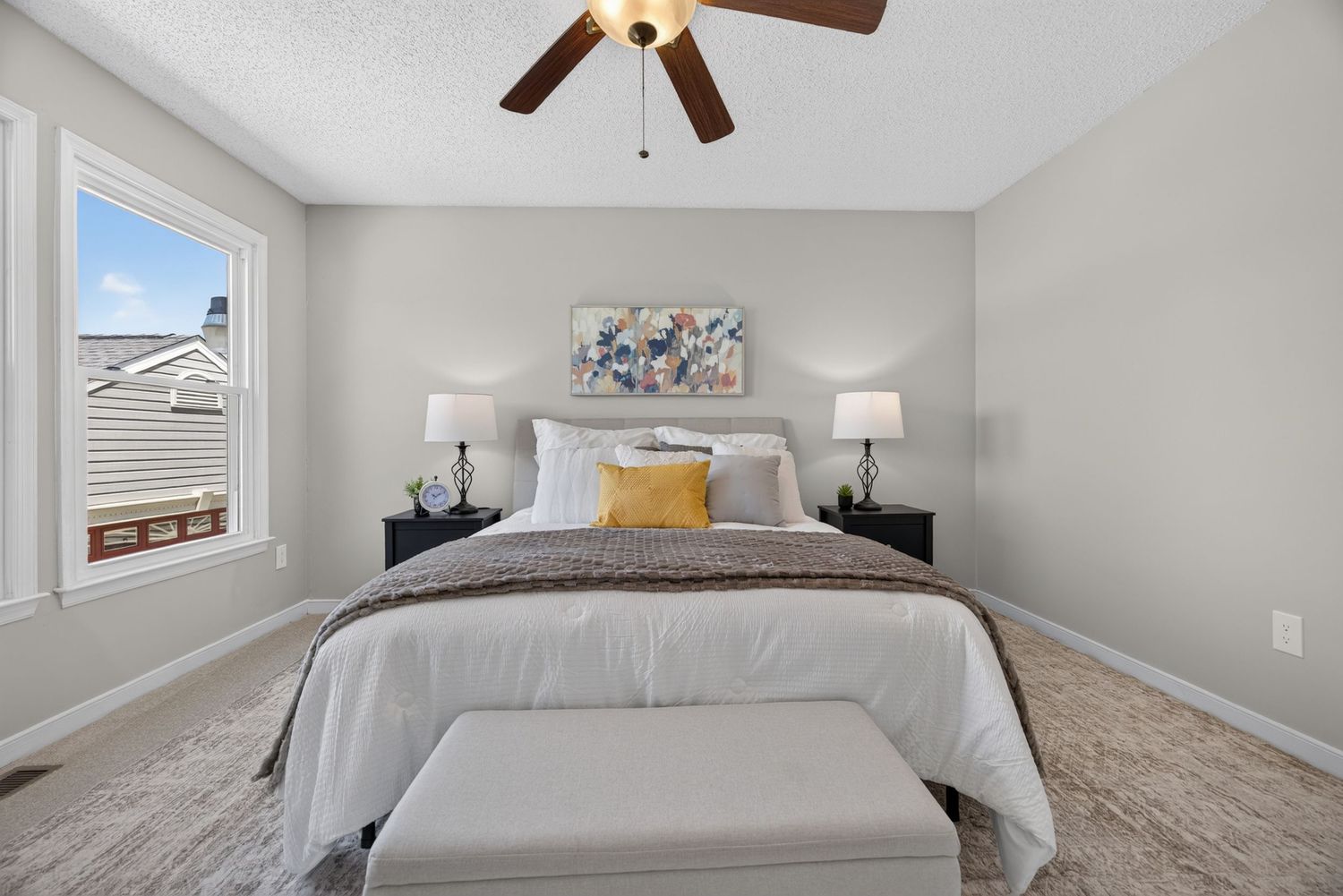 Bright bedroom with a queen bed, yellow accent pillows, ceiling fan, bench, and soft gray walls with carpet flooring.