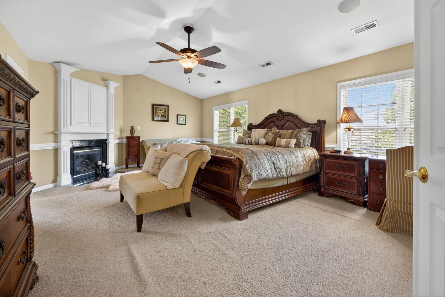 Elegant master bedroom with ornate wood sleigh bed, fireplace, ceiling fan, and plush carpet in neutral tones.