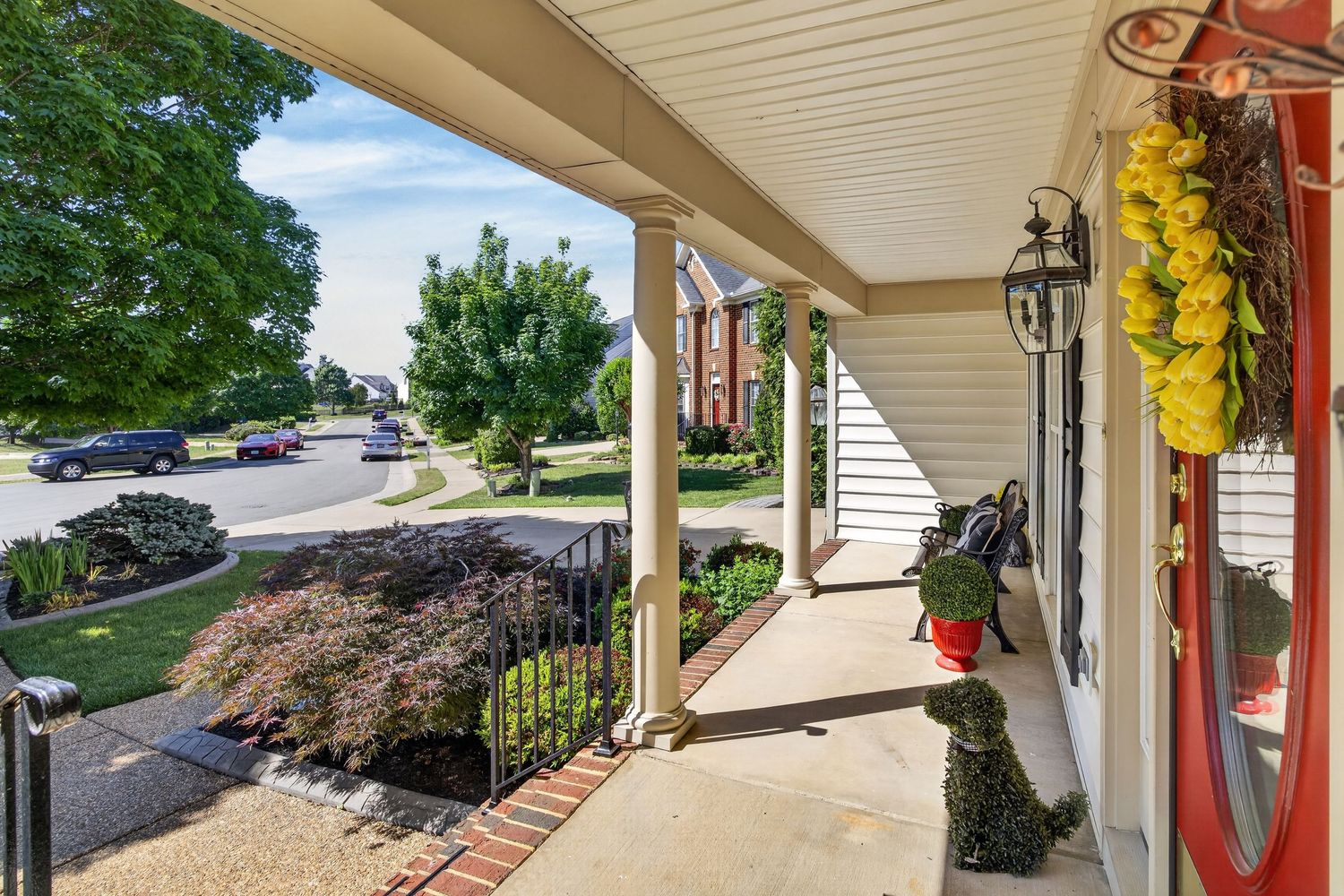 Charming covered front porch with white columns, red door, and lush landscaping overlooking a quiet suburban street.