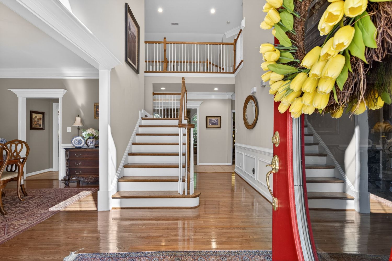 Elegant two-story foyer with hardwood floors, white staircase, wainscoting, and a red front door adorned with yellow tulip wreath.