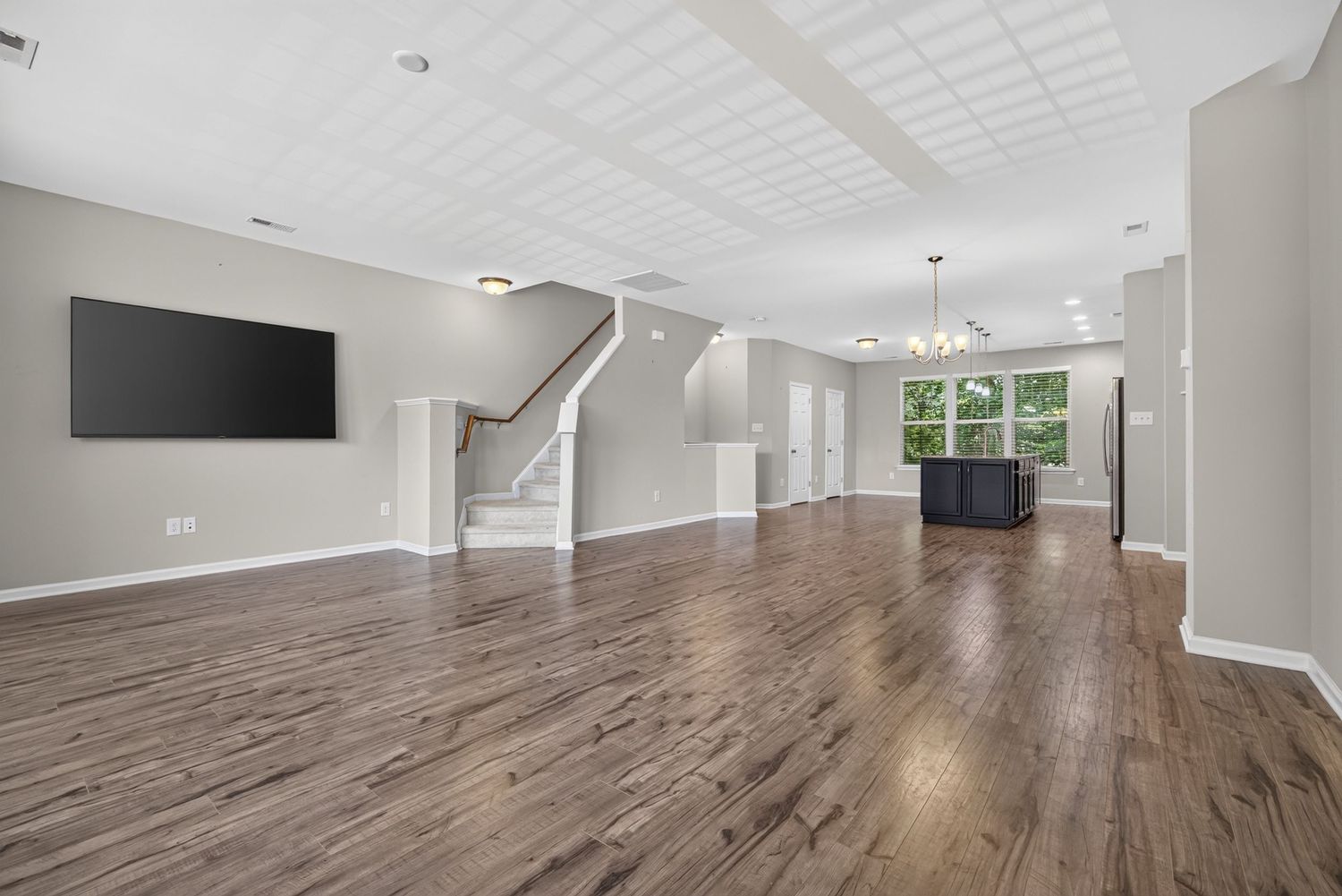 Spacious open-concept living room with dark hardwood floors, wall-mounted TV, staircase, and chandelier-lit dining area.
