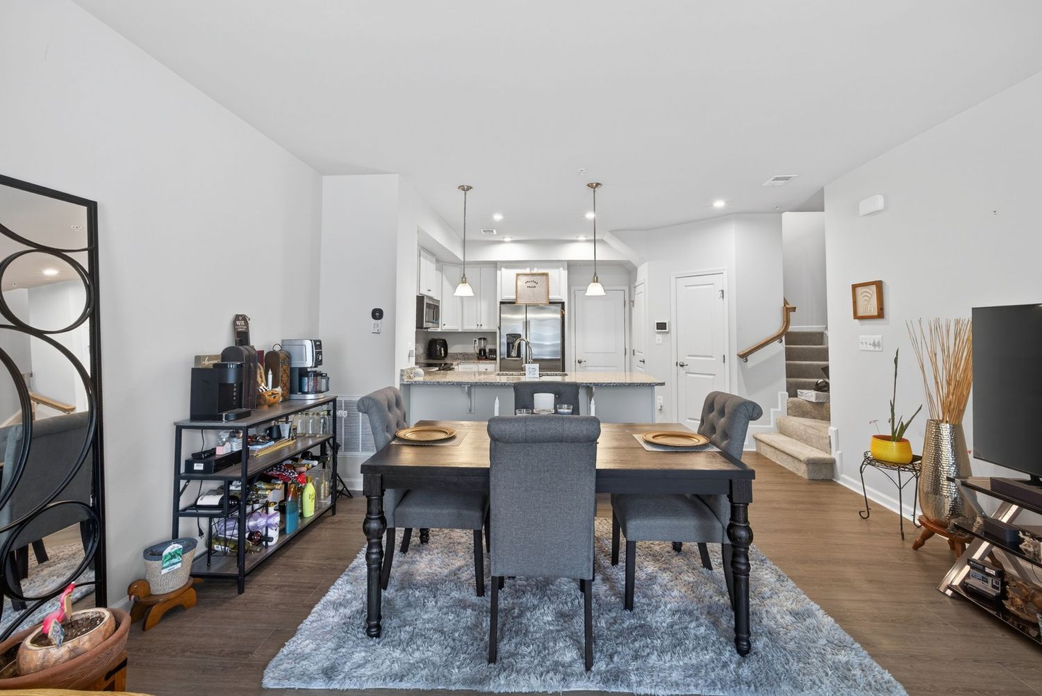 Modern open-concept dining room with gray chairs, wood table, blue rug, and white kitchen with pendant lights in background.