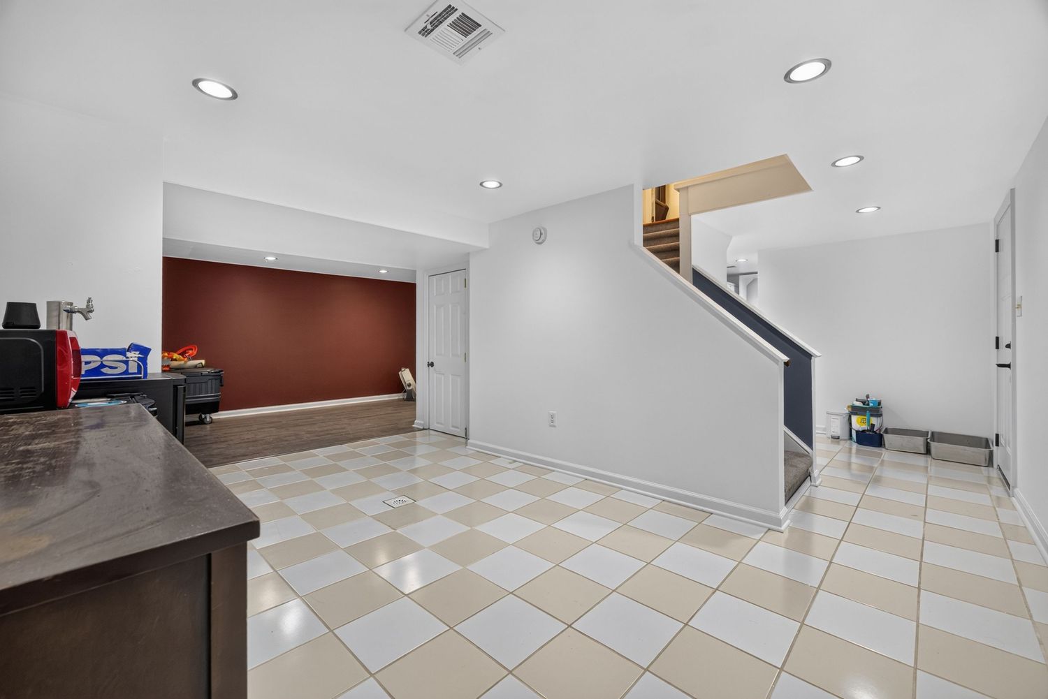Spacious living area with checkered tile floors, recessed lighting, a brown accent wall, and a staircase in the background.
