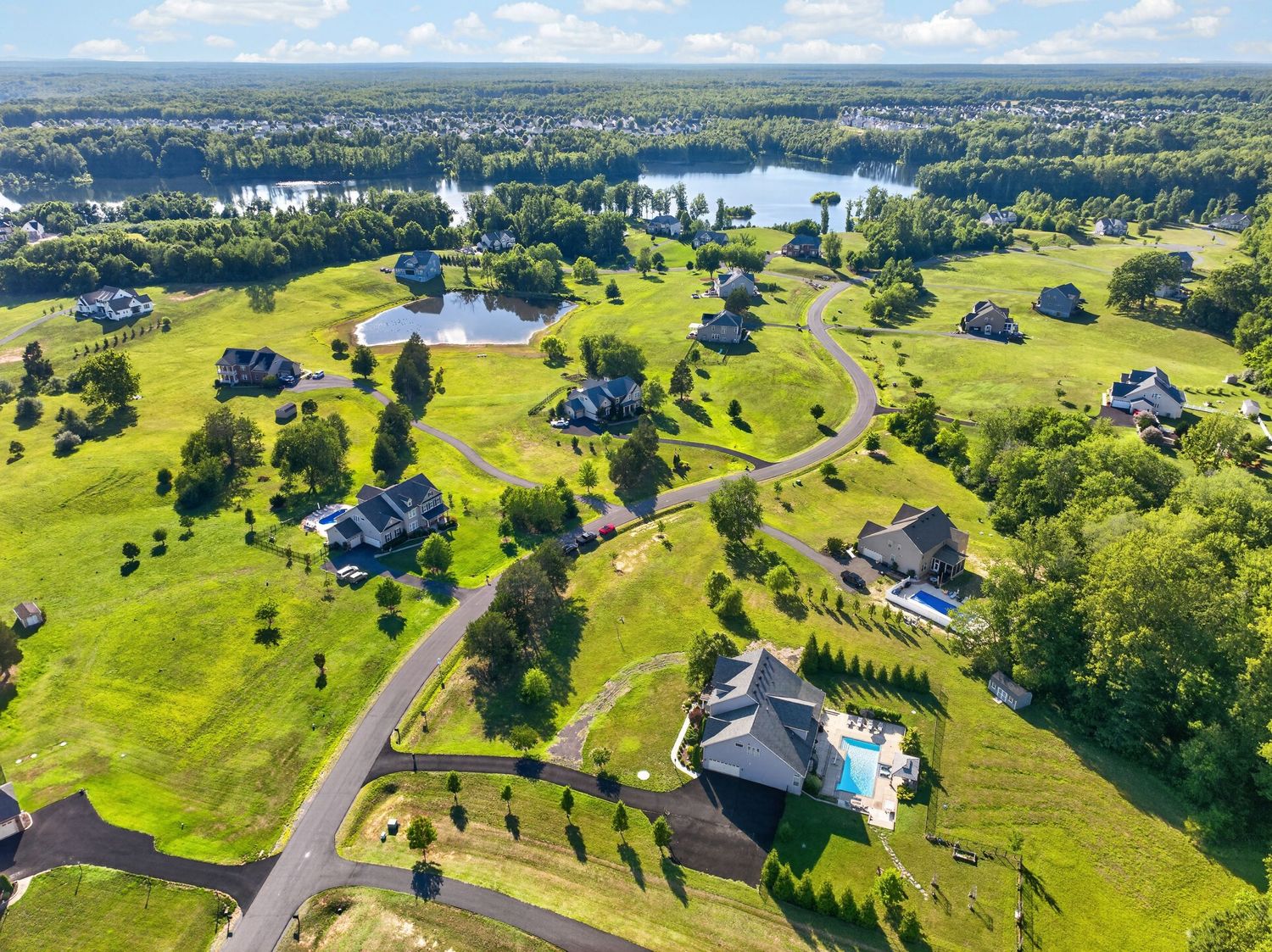 Aerial view of a lush green suburban neighborhood with winding roads, luxury homes, and a scenic lake in the background.
