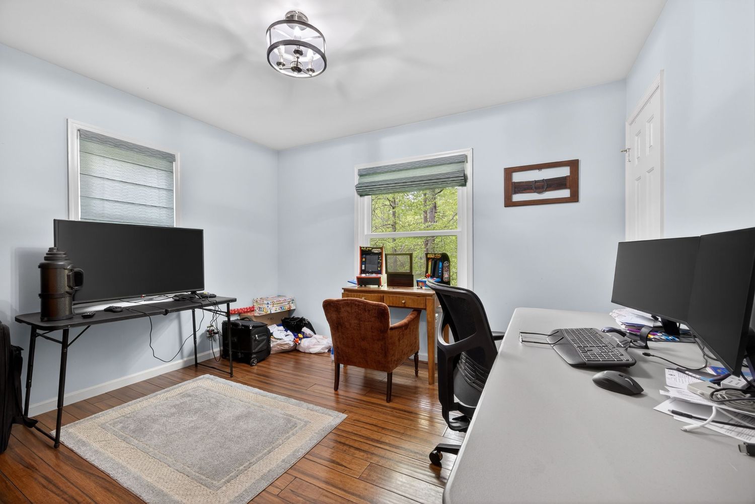 Home office with hardwood floors, two desks, dual monitors, a chandelier, and natural light from two windows.