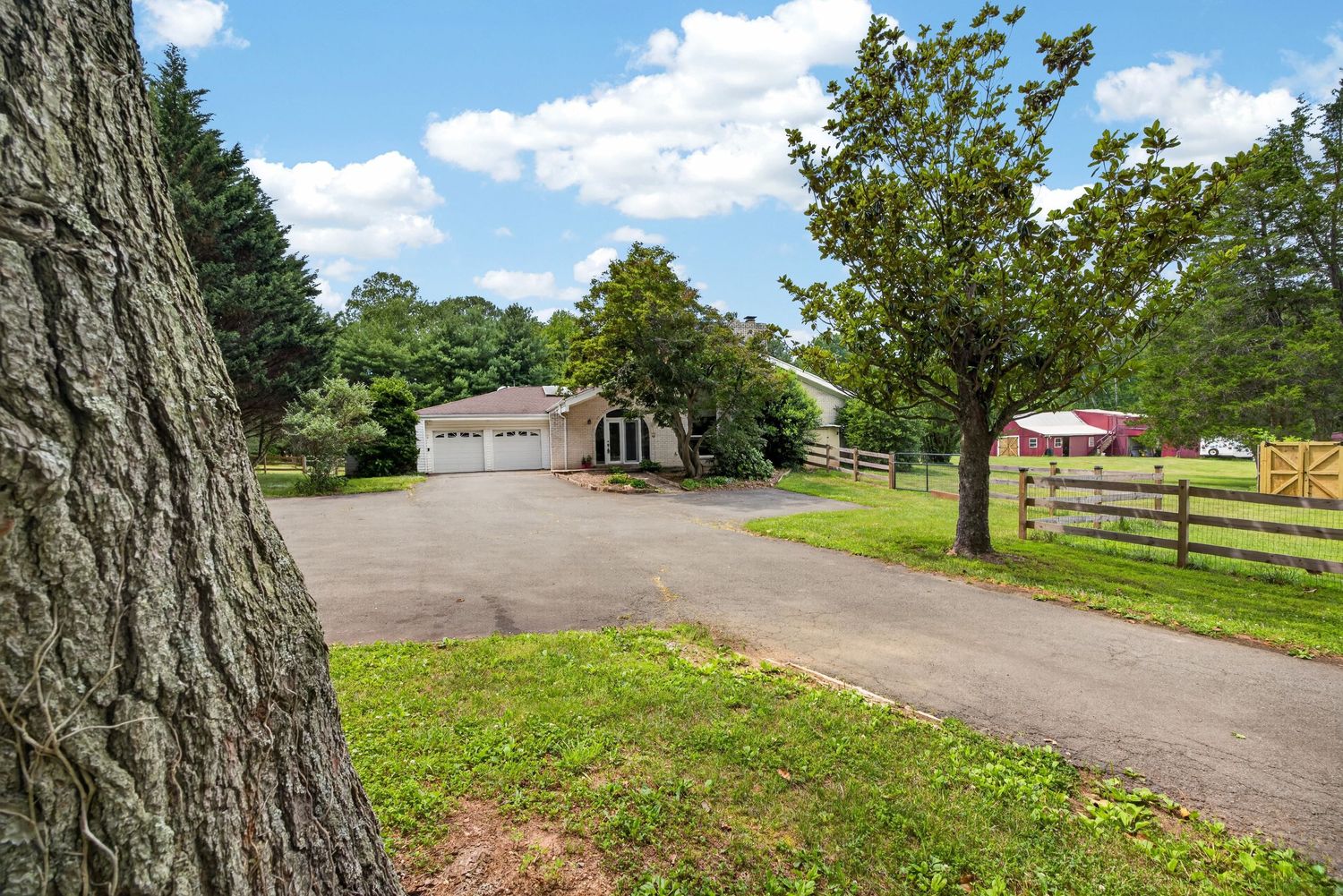 Rural property with circular driveway, detached garage, lush green lawn, mature trees, and wooden fence under a partly cloudy sky.