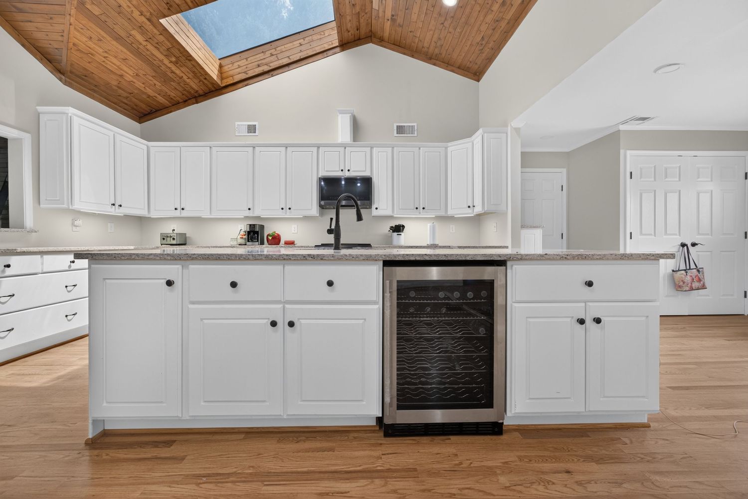 White kitchen with wine fridge, vaulted wood ceiling, skylight, and hardwood floors.