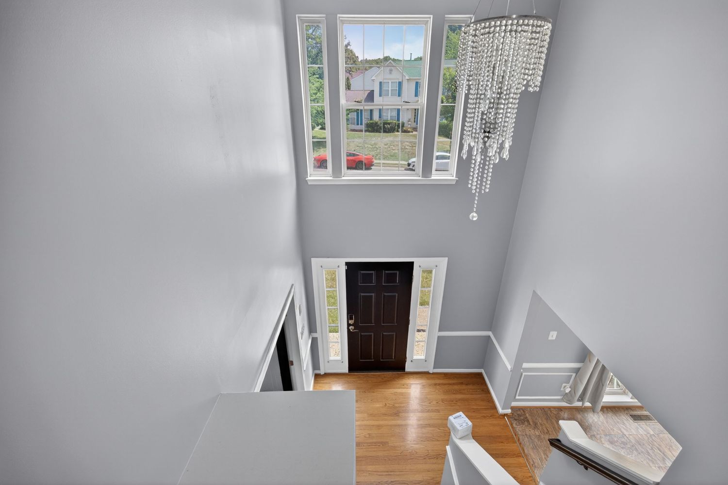 Two-story foyer with hardwood floors, crystal chandelier, dark front door, sidelights, and tall windows overlooking a neighborhood.