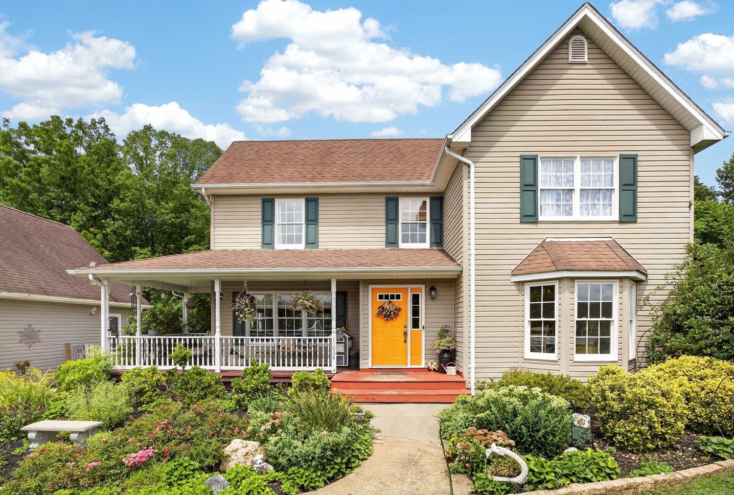 Charming two-story beige colonial home with green shutters, covered porch, orange door, and lush landscaped gardens.