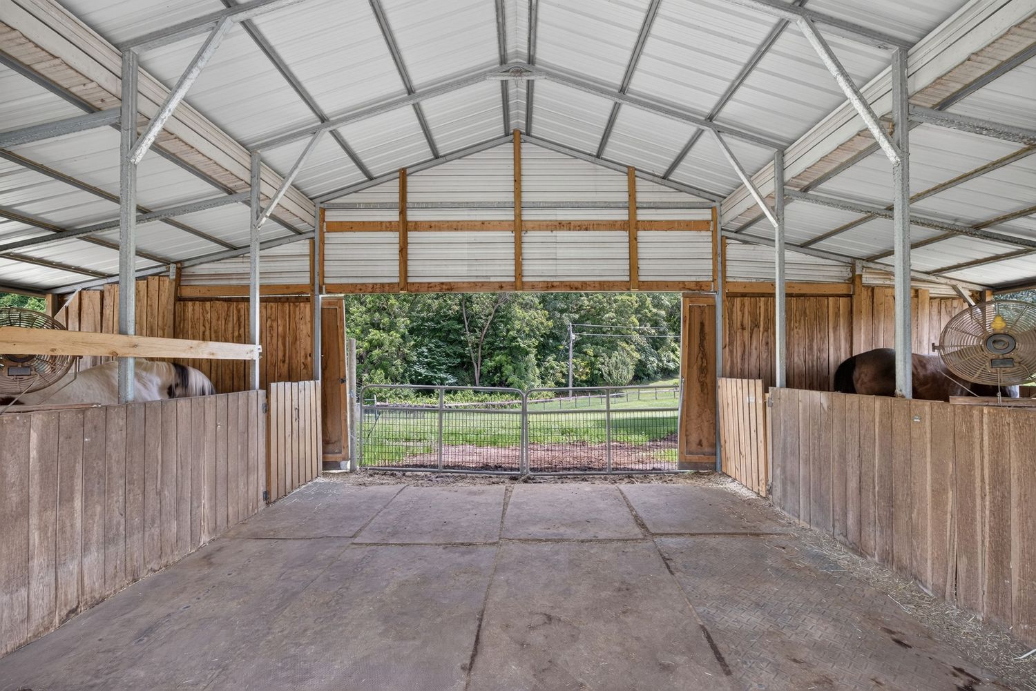 Interior view of an open wooden horse barn with metal roof, concrete floor, and green pasture visible through the open gate.