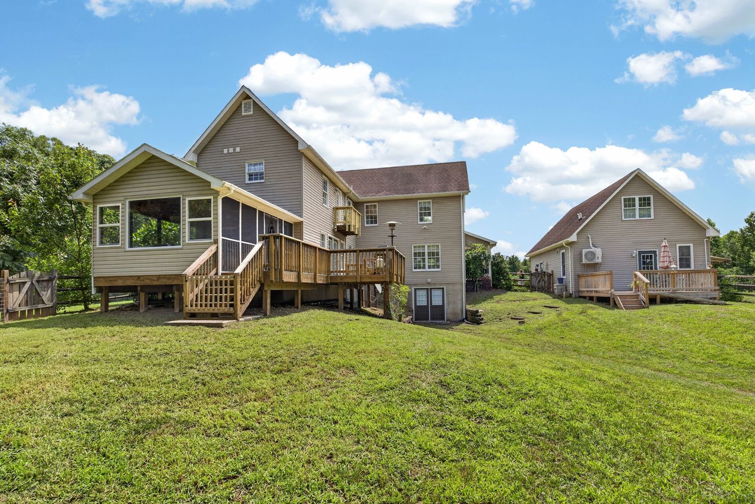 Large two-story home with wood deck, screened porch, and detached garage surrounded by lush green lawn under blue sky.