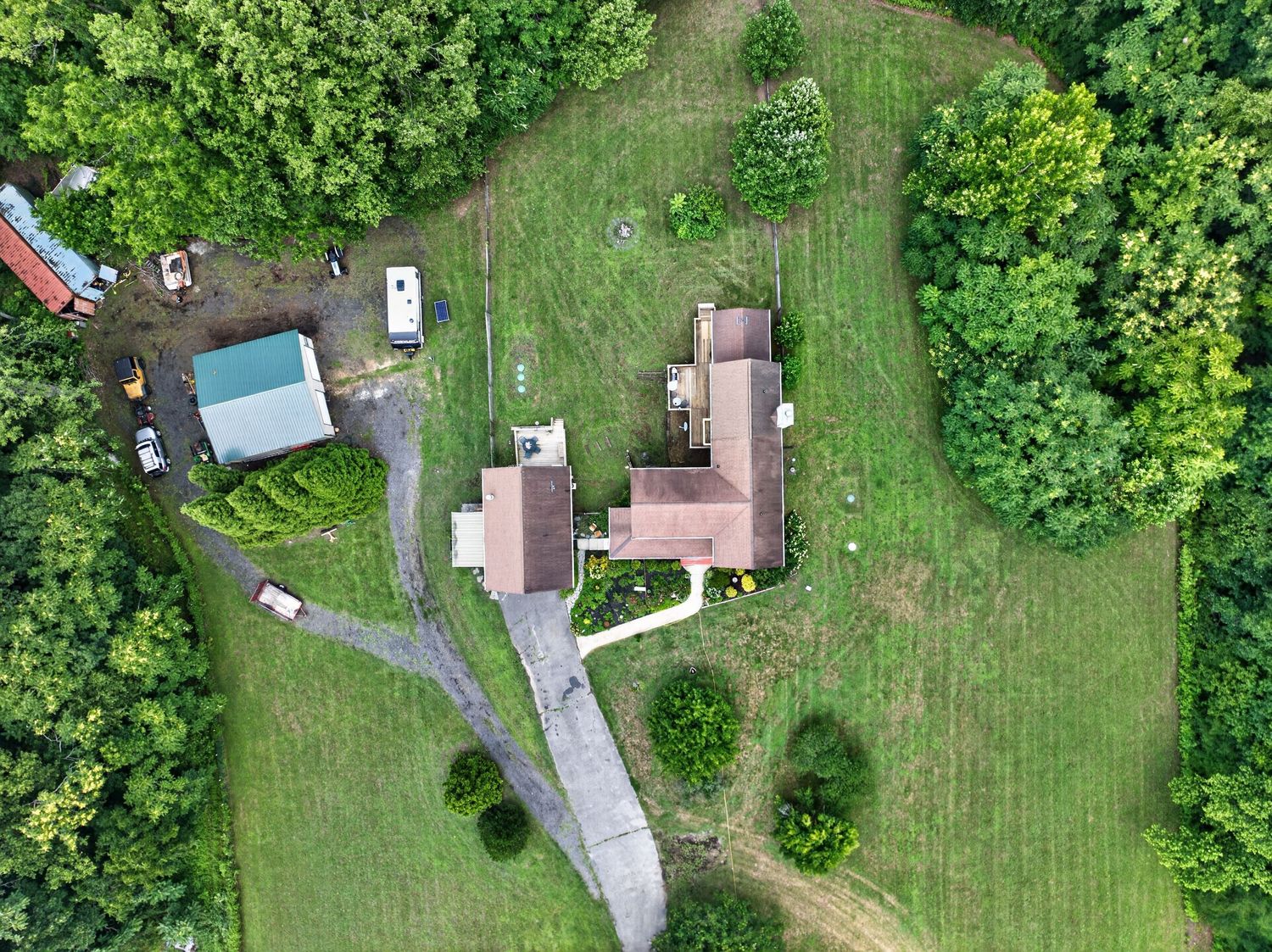 Aerial drone view of a rural property with a house, blue barn, driveway, and lush green surrounding trees.