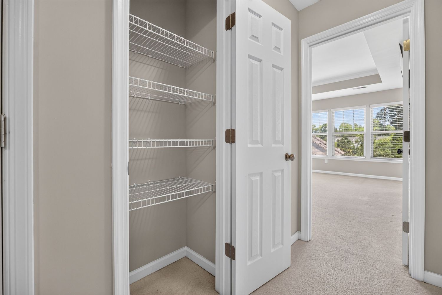 Walk-in closet with white wire shelving beside an open door leading to a bright carpeted bedroom with large windows.