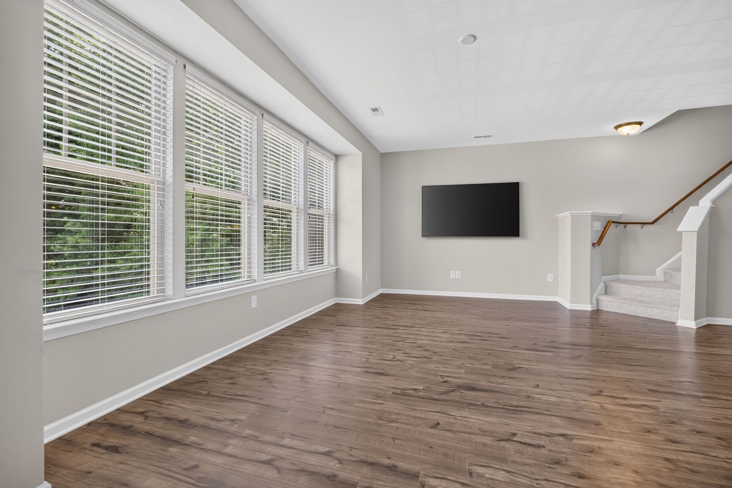 Bright open living room with hardwood floors, large windows with white blinds, wall-mounted TV, and staircase.