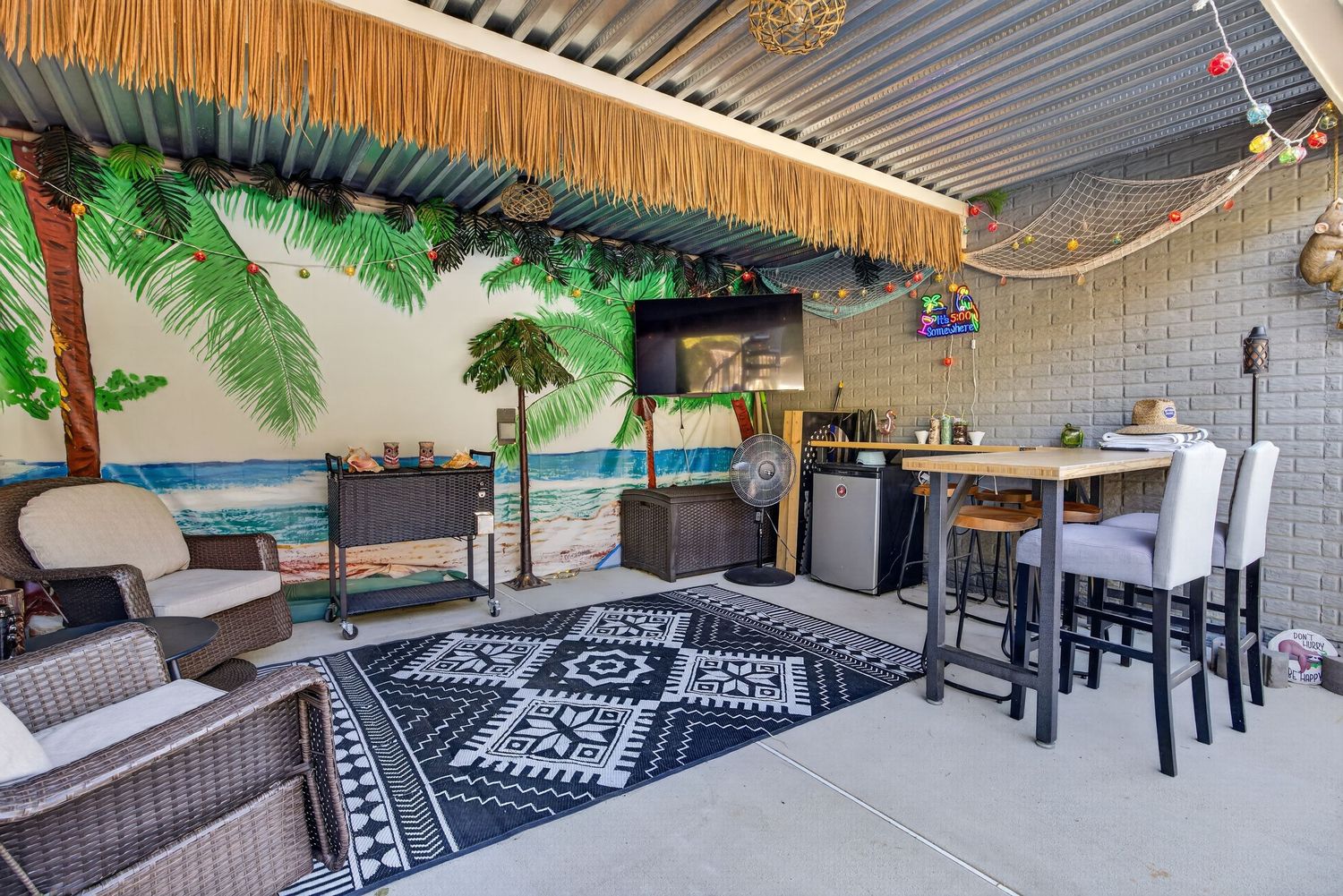 Tropical-themed covered patio with tiki bar, wicker seating, black patterned rug, bar stools, and palm tree mural.