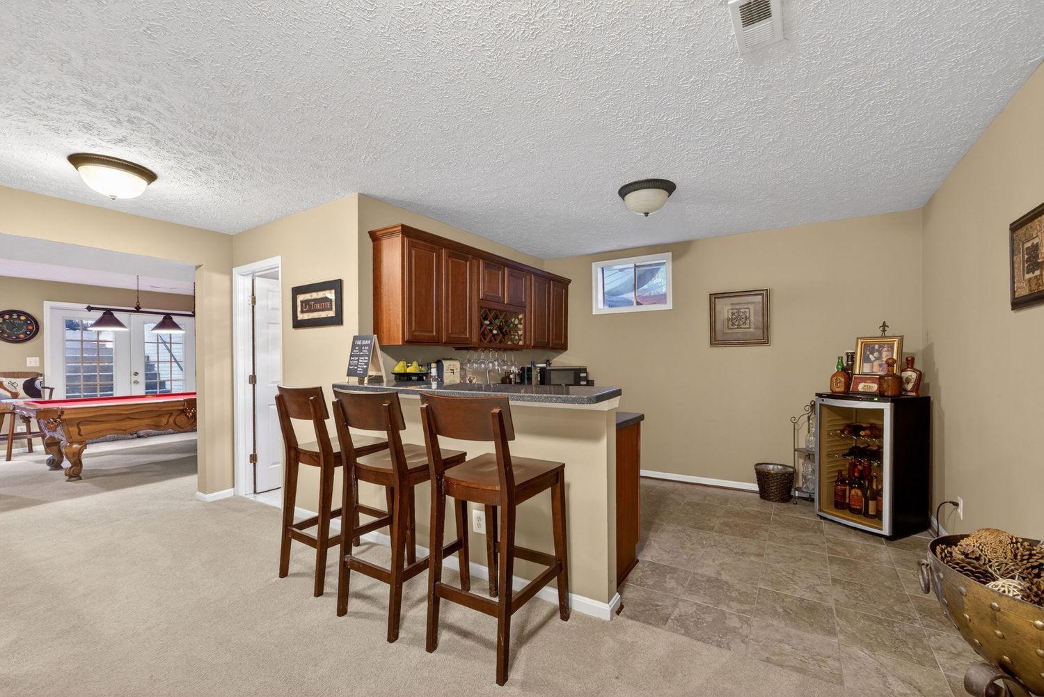Basement bar area with wooden cabinets, bar stools, carpet flooring, and a pool table visible in the background.