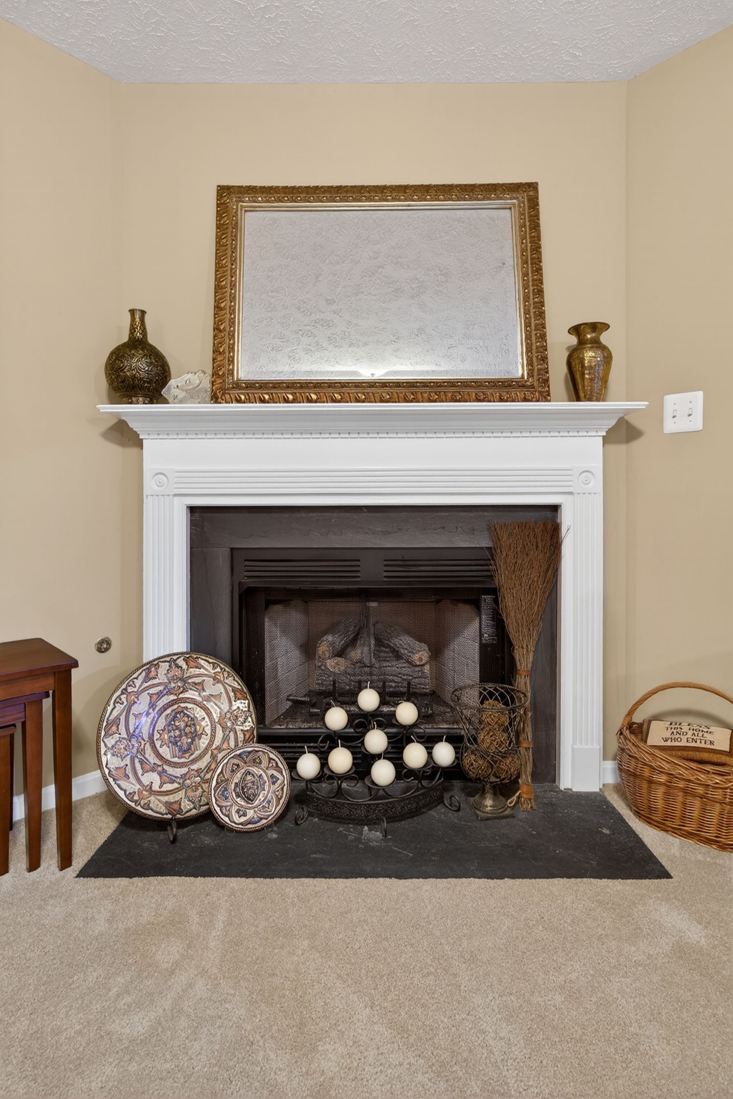 White fireplace mantel with gold mirror, decorative plates, candles, and wicker basket on beige carpet.