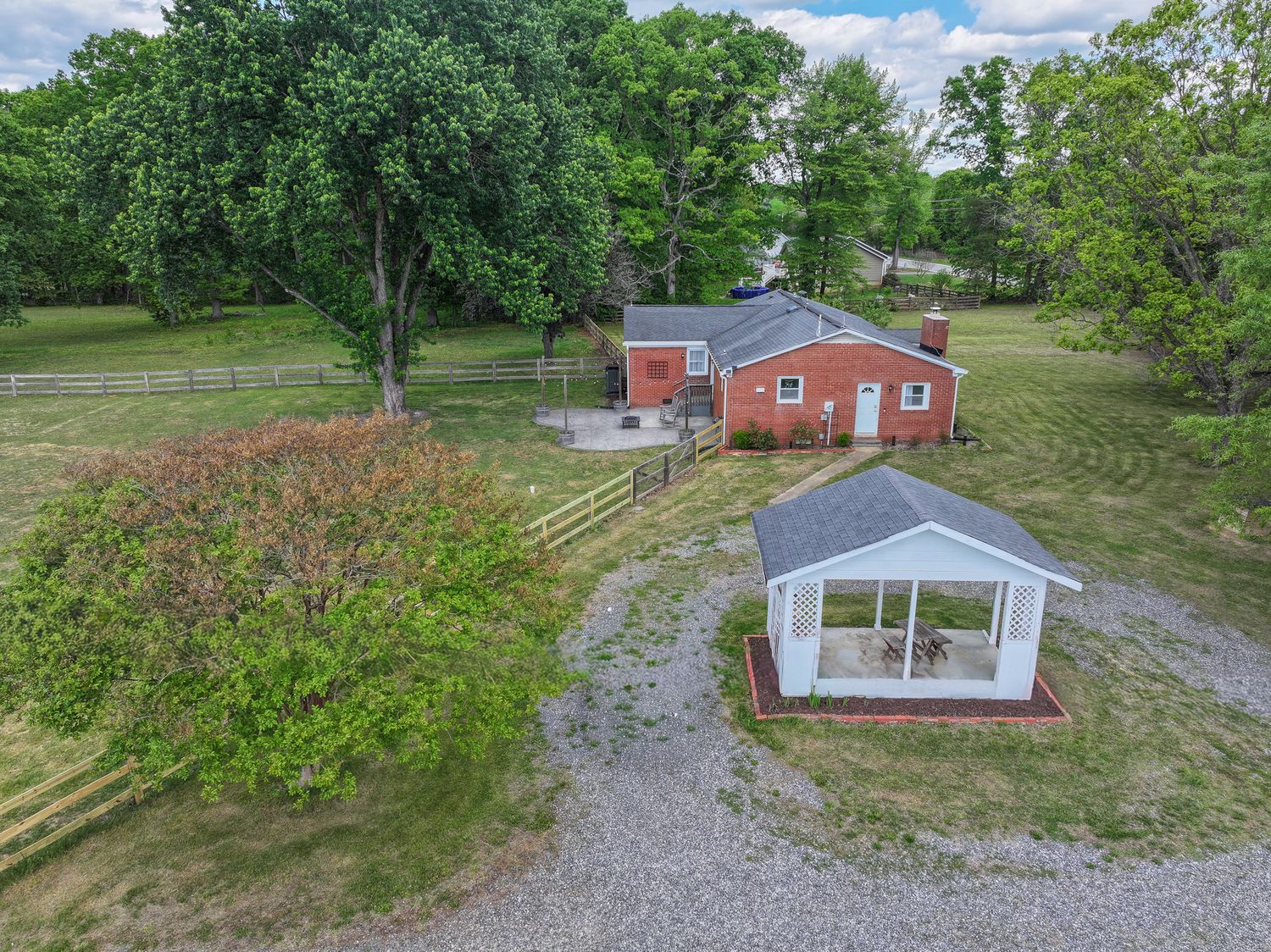 Aerial view of a red brick ranch home with a white greenhouse, gravel driveway, and lush green trees on a spacious rural property.