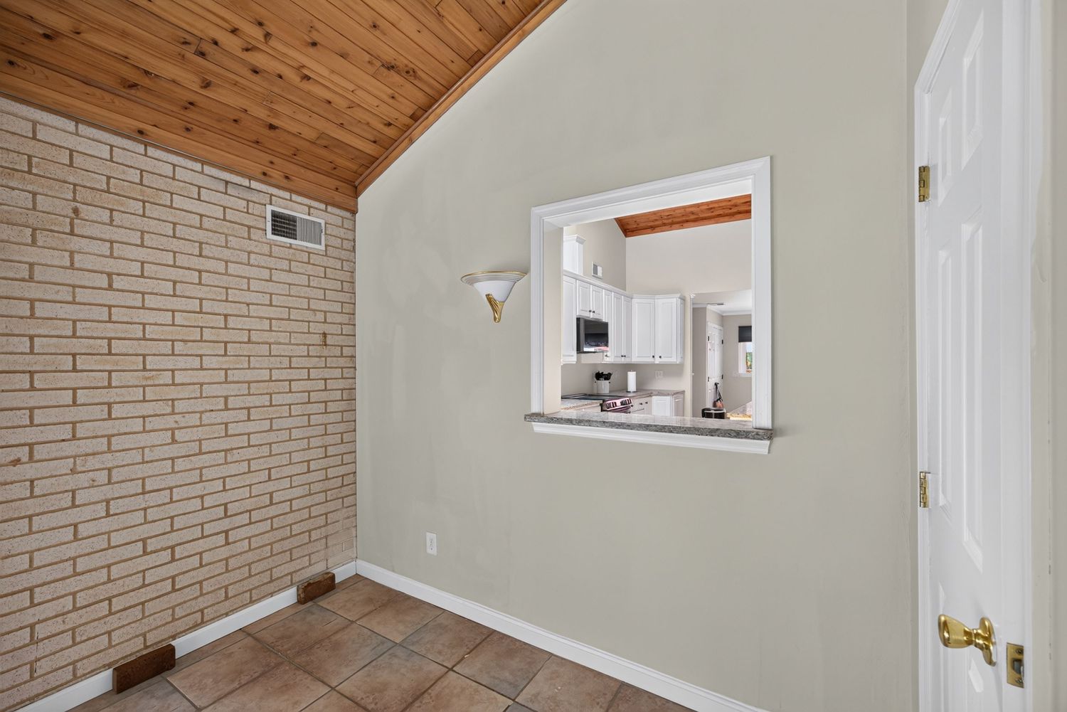 Cozy room with brick accent wall, wood plank ceiling, tile floor, and pass-through window to bright kitchen.