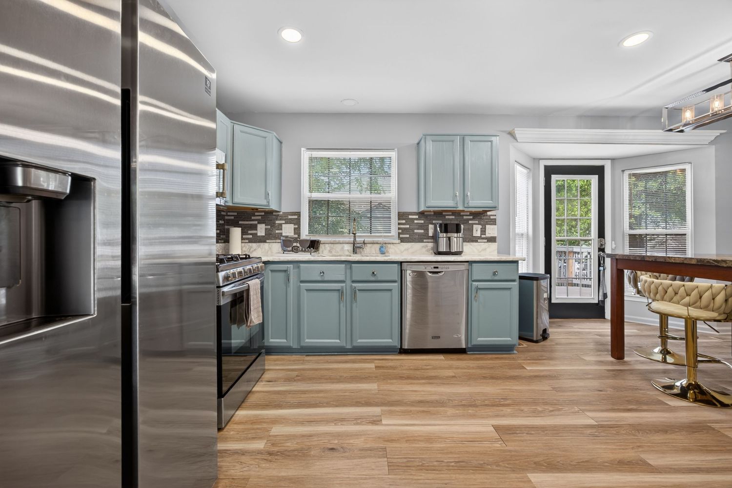 Modern kitchen with teal cabinets, stainless steel appliances, light wood floors, and natural light from multiple windows.