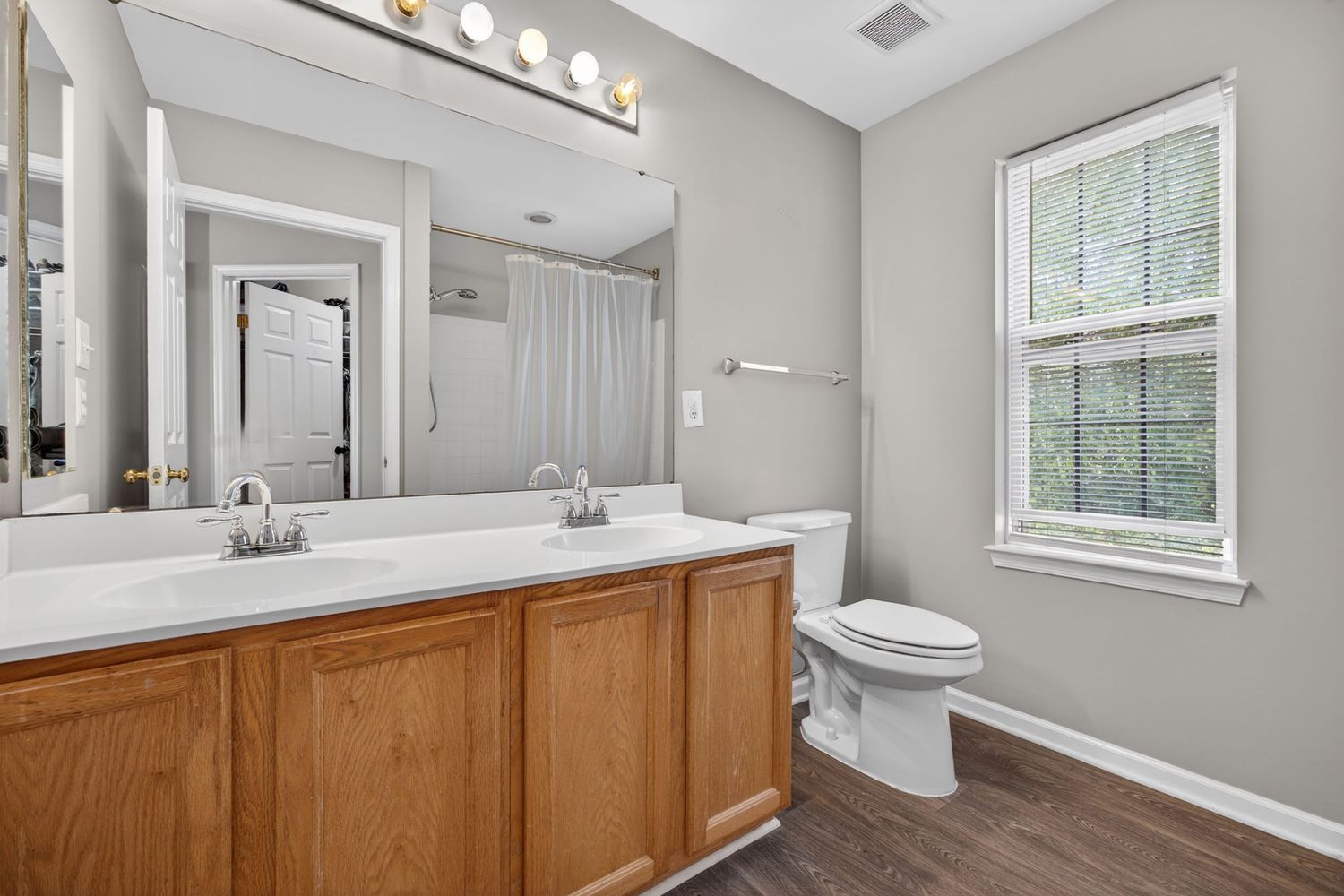 Modern bathroom with double vanity, wood cabinets, large mirror, white countertop, toilet, and hardwood floors.