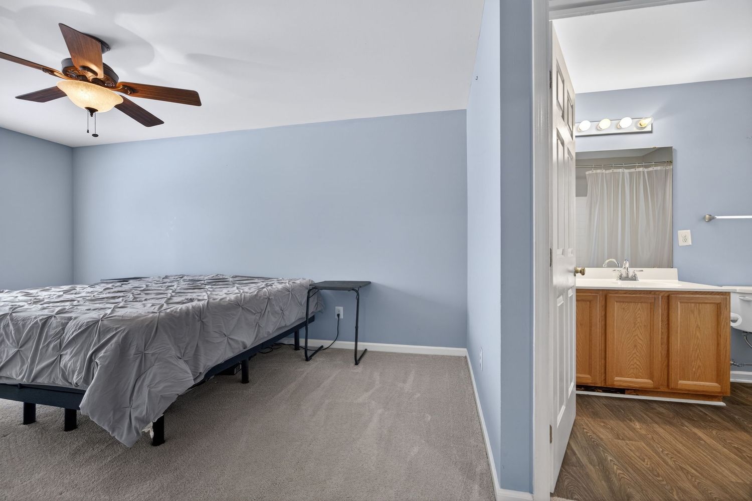 Bedroom with blue walls, ceiling fan, gray bed, and en-suite bathroom with wooden vanity visible through doorway.