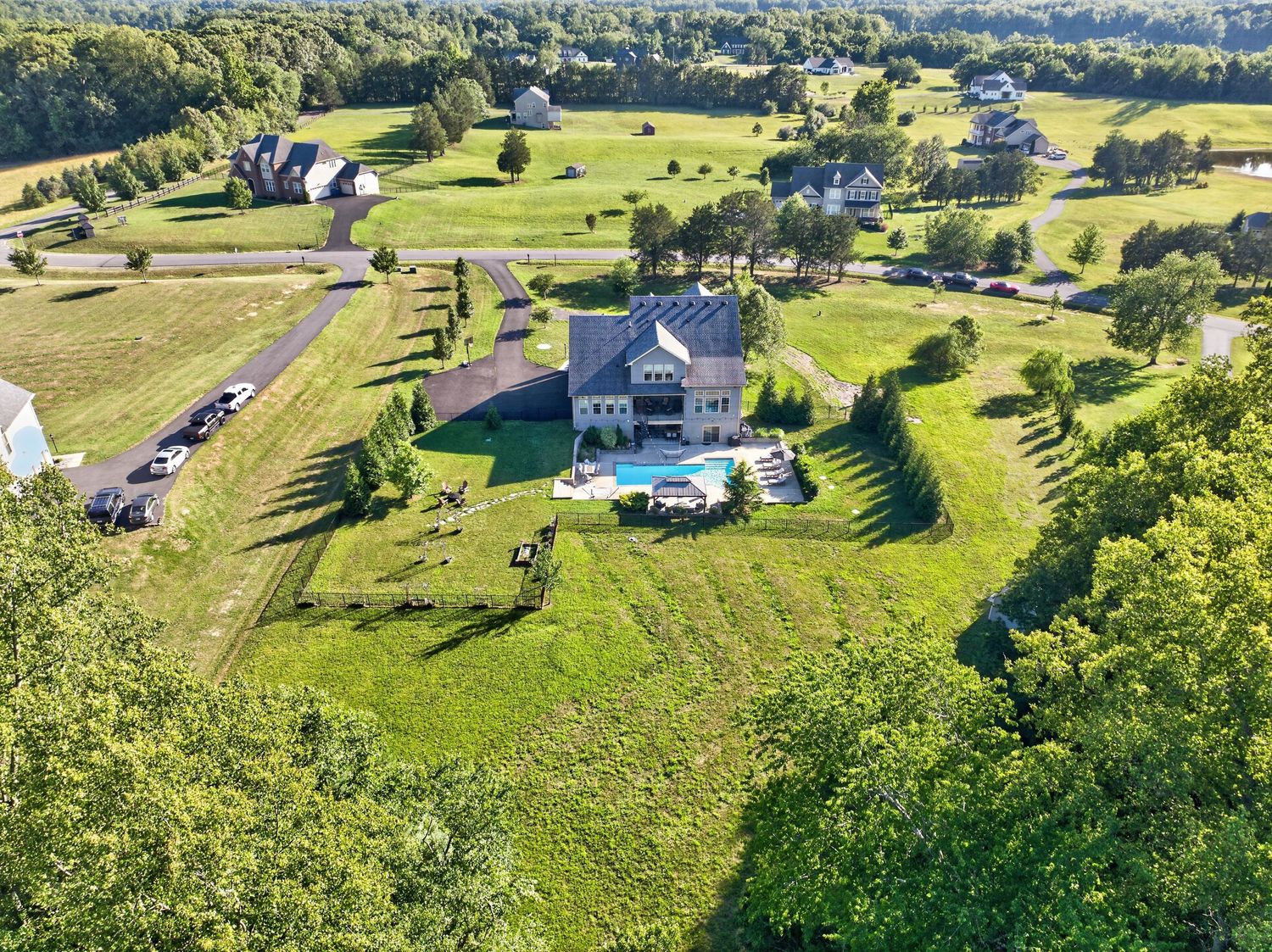 Aerial view of a large suburban home with a pool surrounded by lush green lawns and trees in a peaceful neighborhood.