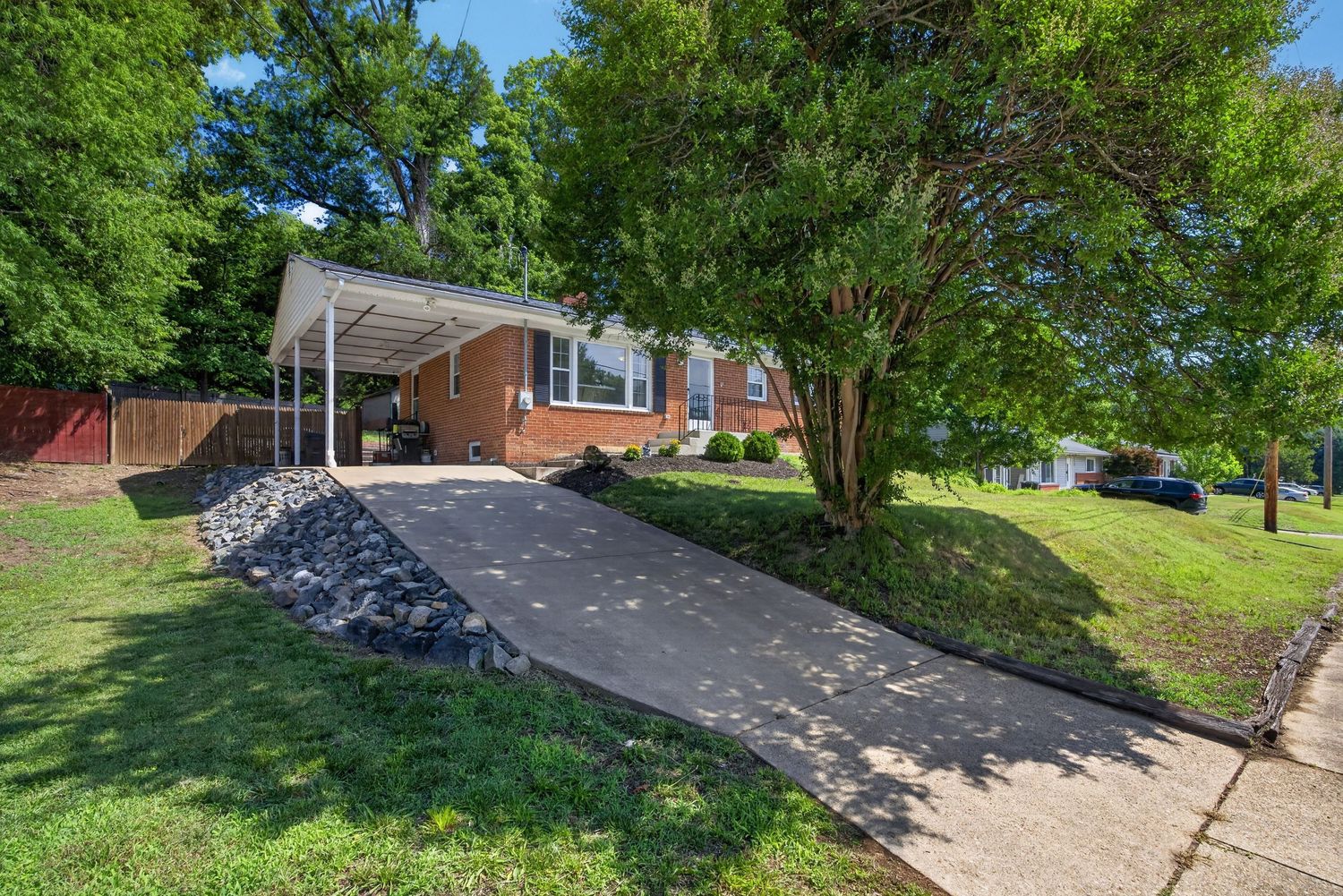 Brick ranch home with carport, concrete driveway, rock landscaping, and lush green lawn surrounded by mature trees.
