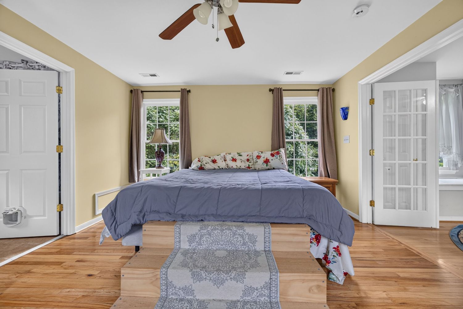 Bright bedroom with hardwood floors, ceiling fan, blue bedspread, area rug, and multiple windows allowing natural light.