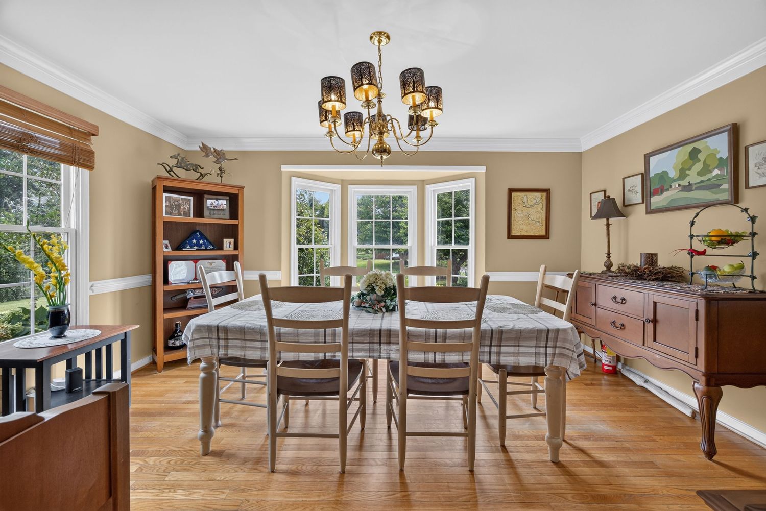 Elegant dining room with chandelier, plaid tablecloth, hardwood floors, bay window, and wooden sideboard in a warm neutral space.