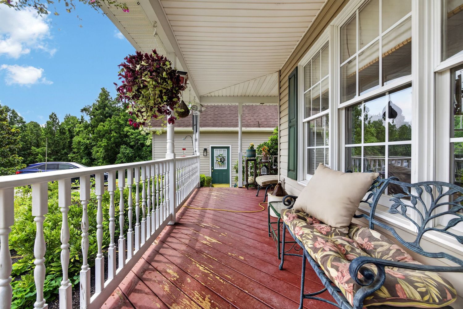 Charming wraparound porch with red deck, white railings, hanging plants, and elegant iron bench with floral cushions.