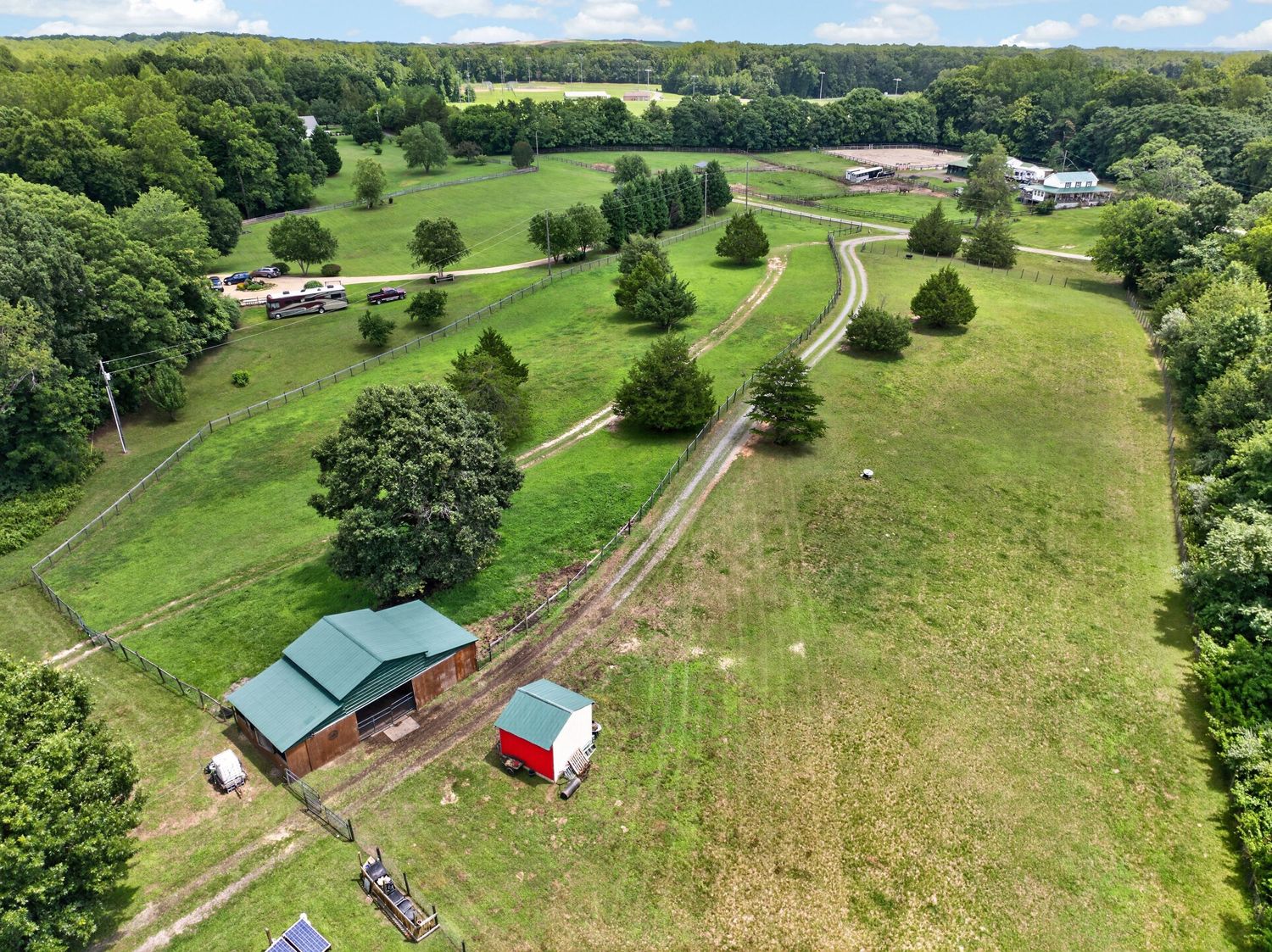 Aerial view of a rural property featuring a red barn with green roof, open green fields, winding driveway, and surrounding woodland.