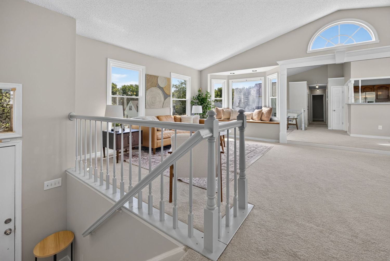 Bright open hallway with white staircase railing, plush carpet, and vaulted ceilings leading to a sunlit living area.