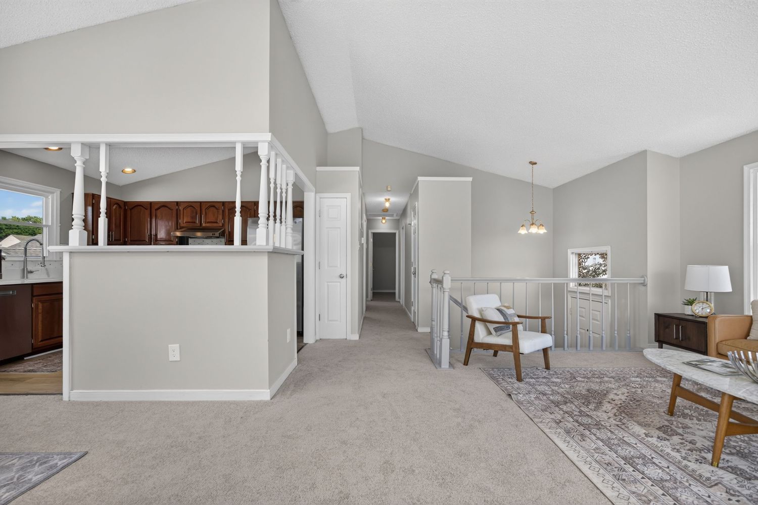Bright open-concept living area with vaulted ceilings, light gray carpet, white railings, and a view into the kitchen.