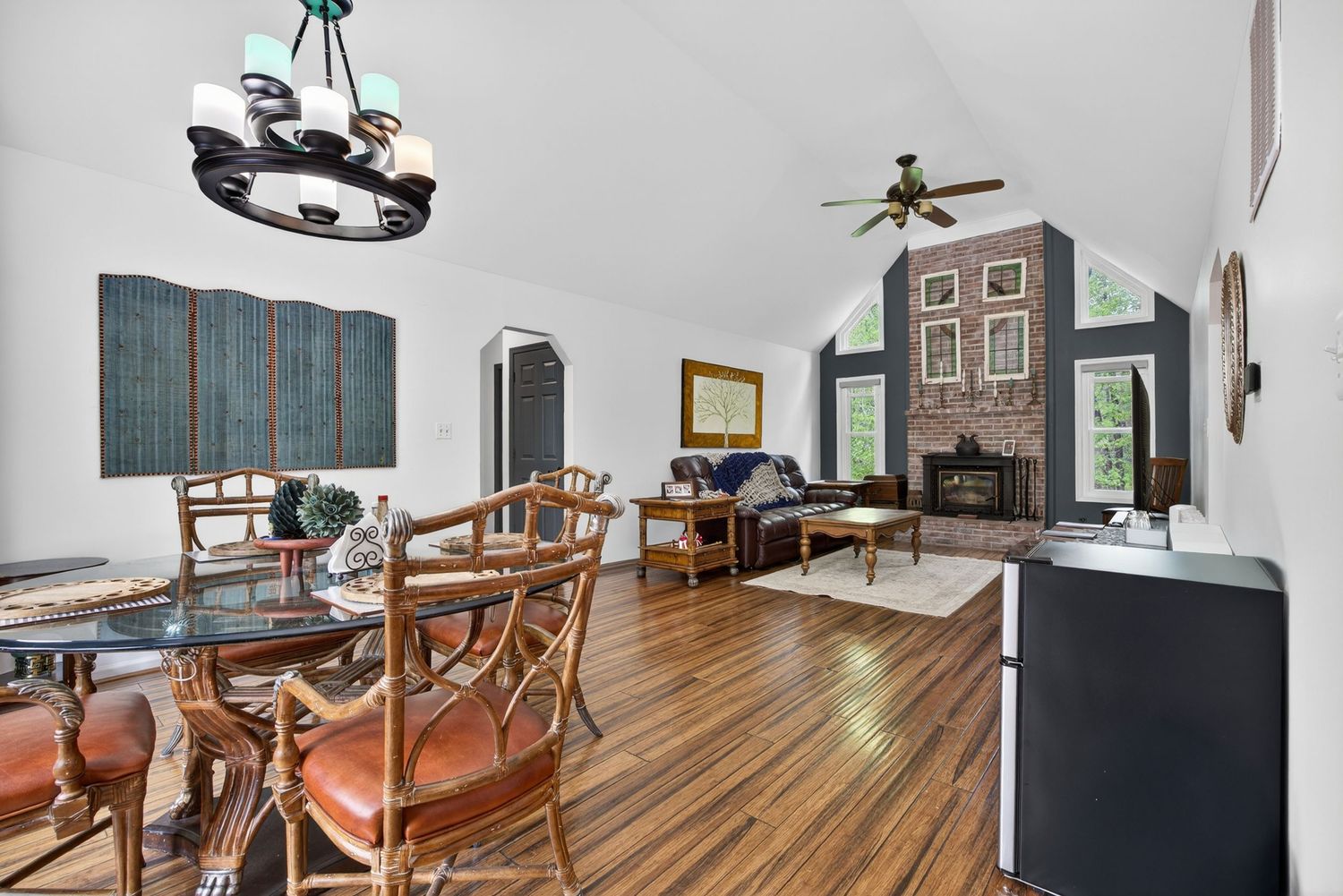 Open-concept living and dining room with hardwood floors, vaulted ceiling, ceiling fan, fireplace, and elegant chandelier.