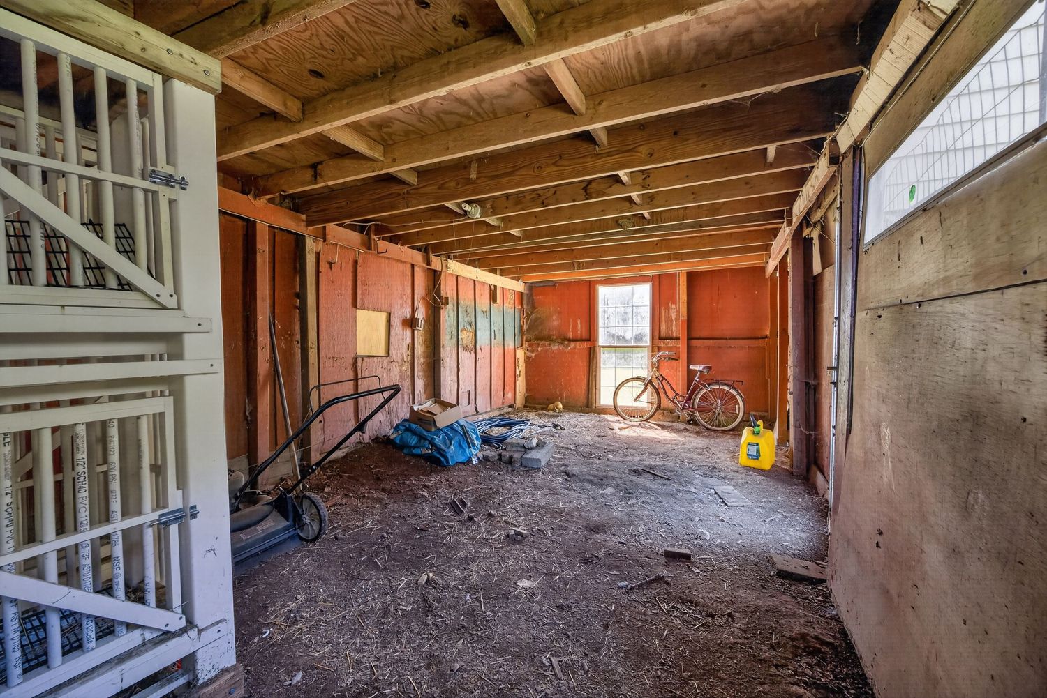 Unfinished garage interior with wooden beam ceiling, orange bicycle, hand truck, and scattered items on dirt floor.
