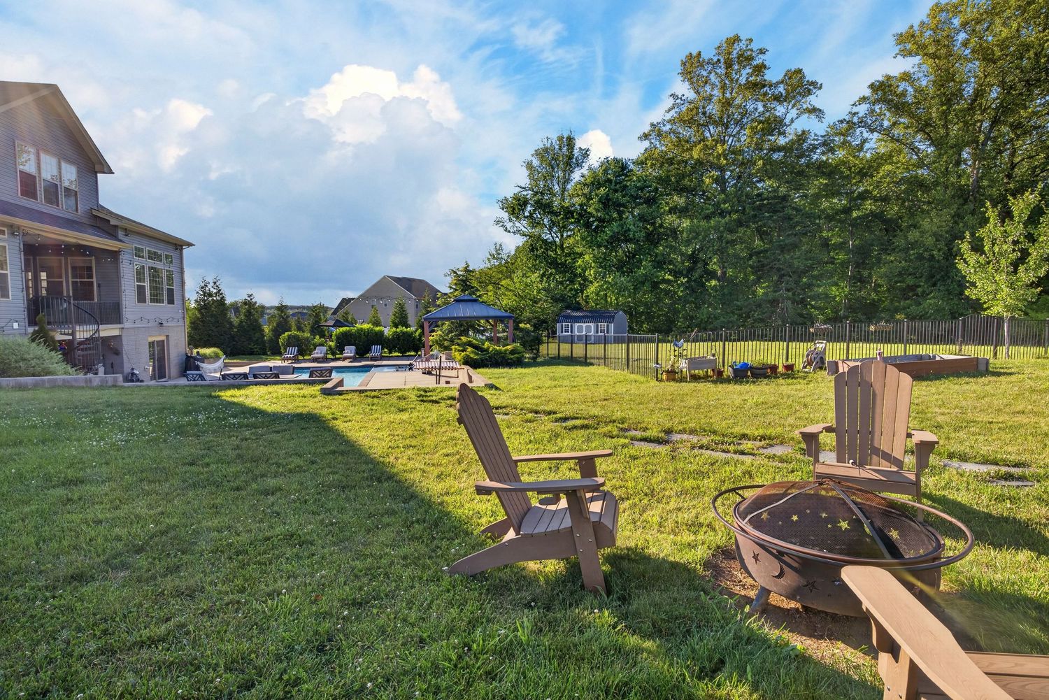 Spacious backyard with Adirondack chairs, fire pit, lush green lawn, pool, and mature trees under a partly cloudy sky.