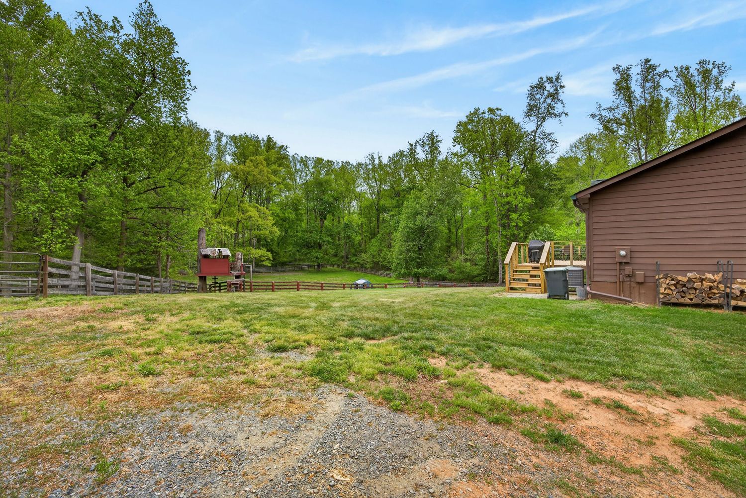 Spacious backyard with lush green trees, wooden fence, gravel driveway, and firewood stacked beside a brown building.