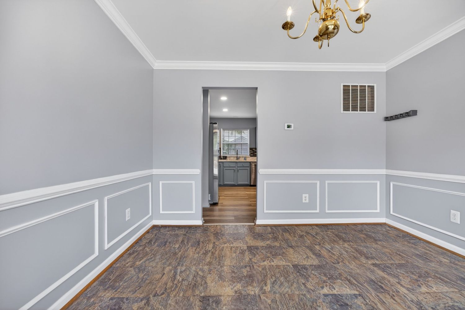 Empty dining room with gray wainscoting, dark hardwood floors, gold chandelier, and open doorway to kitchen.