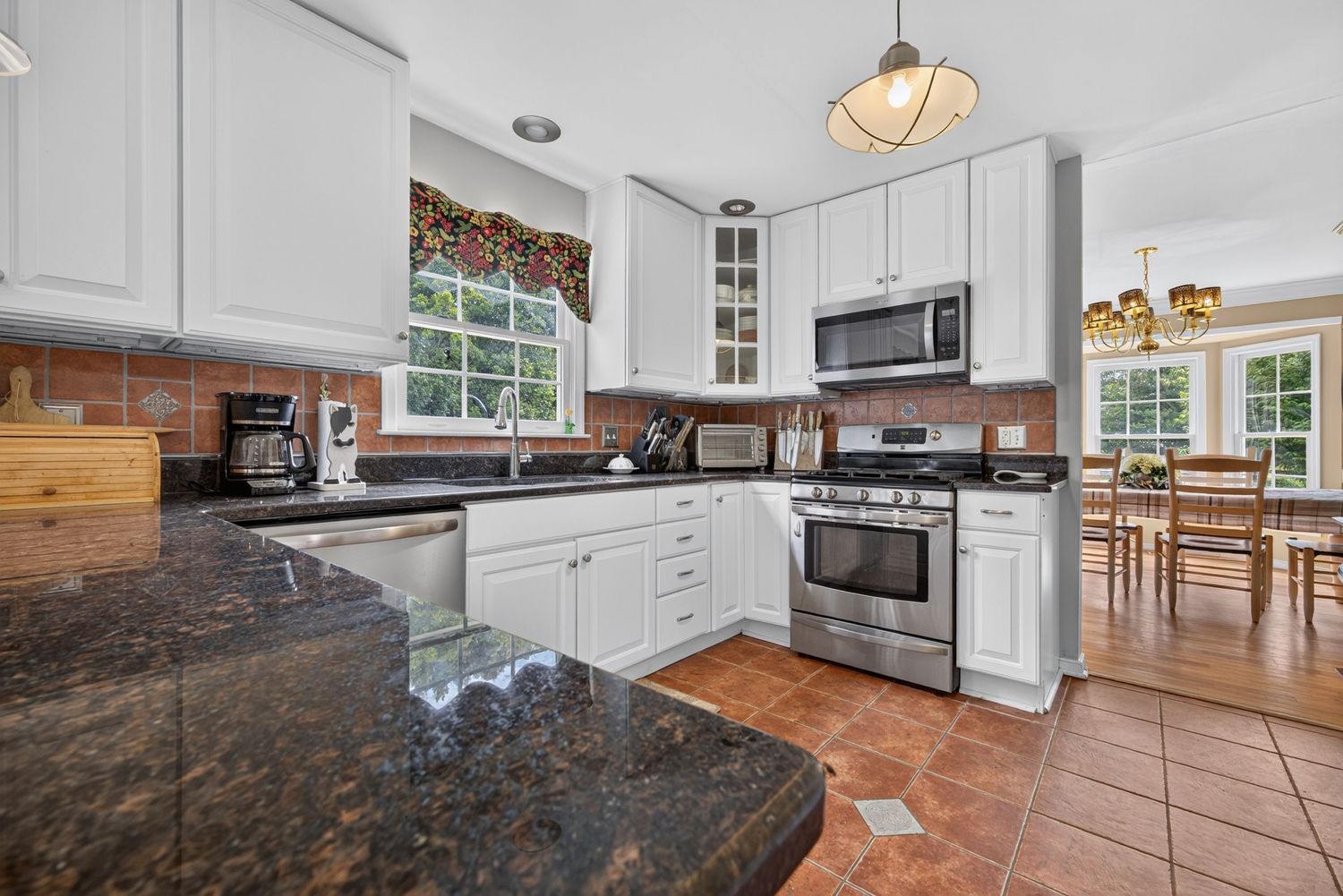 White kitchen with granite countertops, stainless steel appliances, terracotta tile floors, and a dining area visible in the background.