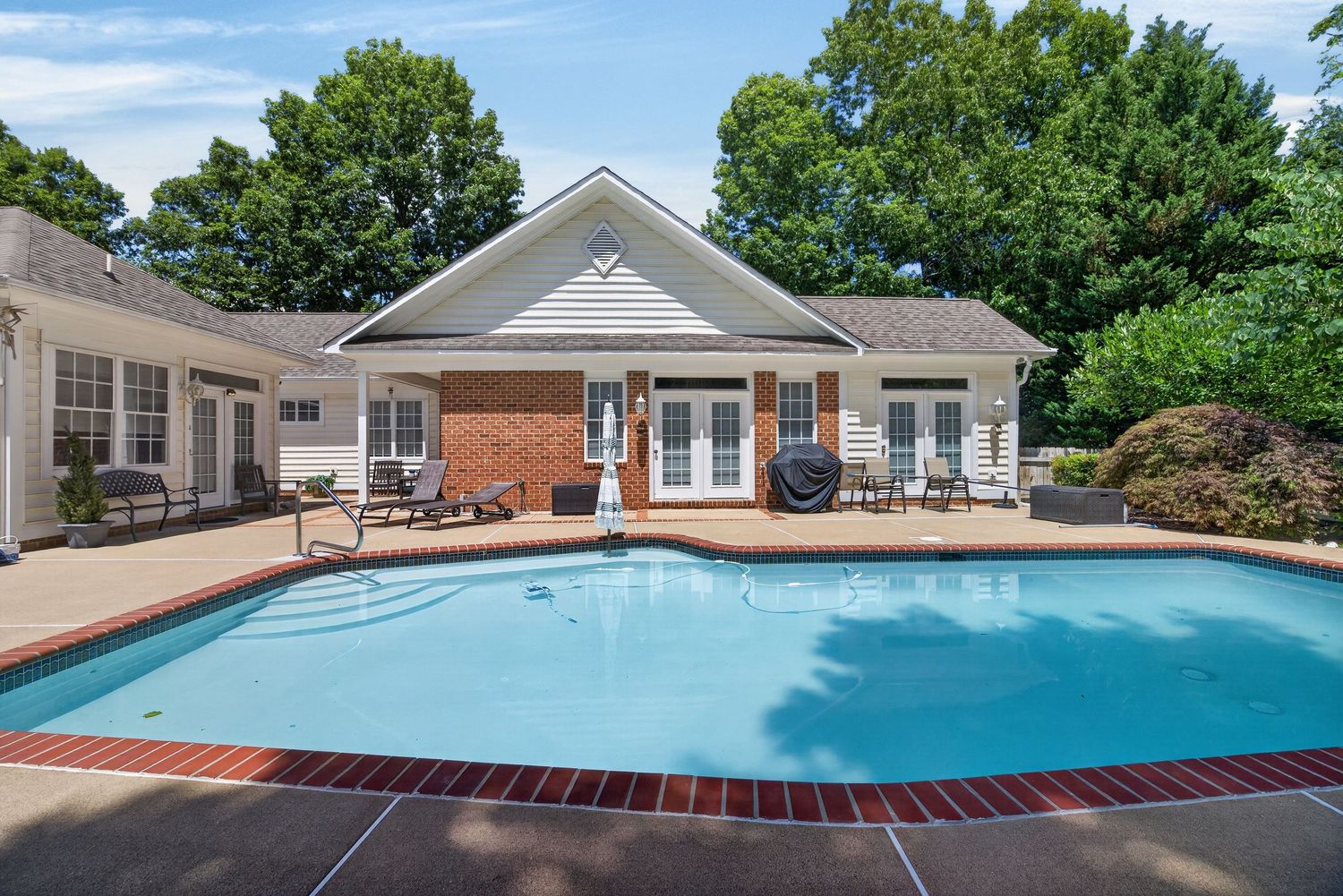 Outdoor pool with brick accents beside a clubhouse with French doors, patio seating, and lush green trees.
