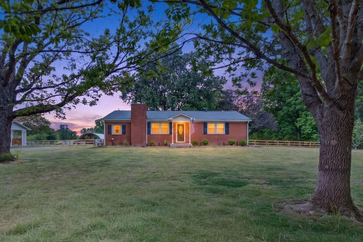 Brick ranch home at dusk with lush green lawn, large shade trees, and warm interior lights glowing at twilight.