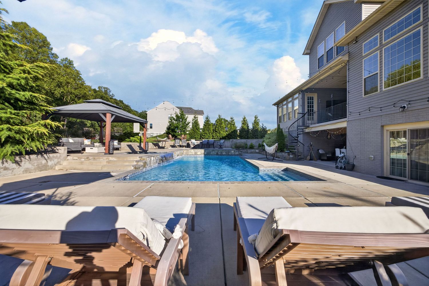Luxury outdoor pool with lounge chairs, stone patio, pergola, and large home backdrop under a partly cloudy sky.